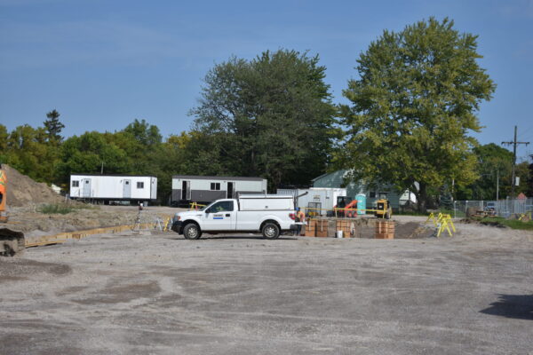 Construction site of future St. Clair Medical Centre. Work trucks and trailers are parked in the background, the ground has been worked/prepped for foundation. Tall green trees in the background.