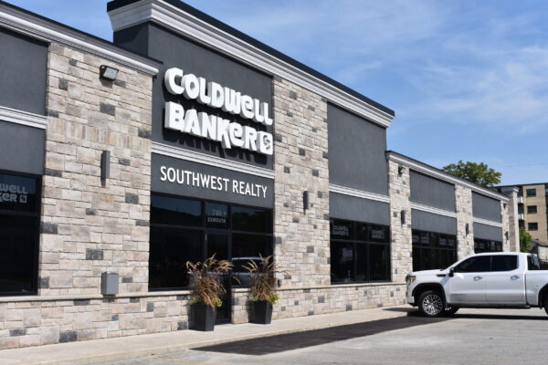 Front left view of the Coldwell Banker Southwest Realty building. One-story commercial building with stone and grey brickwork, tinted windows throughout, and on-site parking in front. The building has a white Coldwell Banker sign on the front.