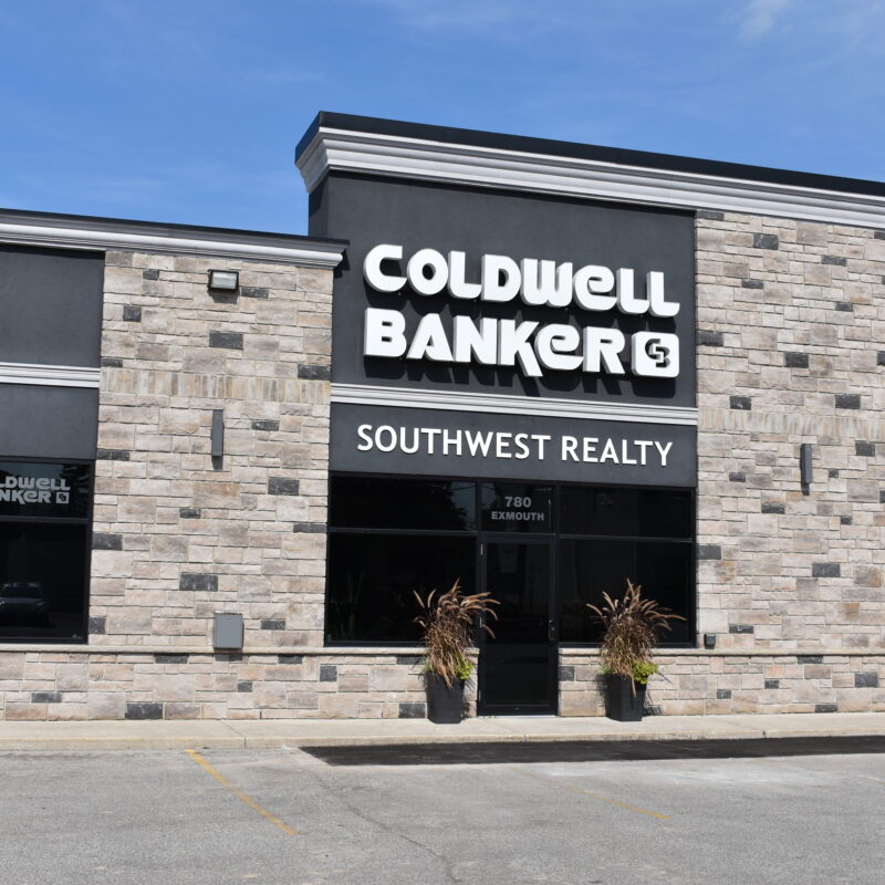 Front view of the Coldwell Banker Southwest Realty building. One-story commercial building with stone and grey brickwork, tinted windows throughout, and on-site parking in front. The building has a white Coldwell Banker sign on the front and a smaller Southwest Realty sign below that.