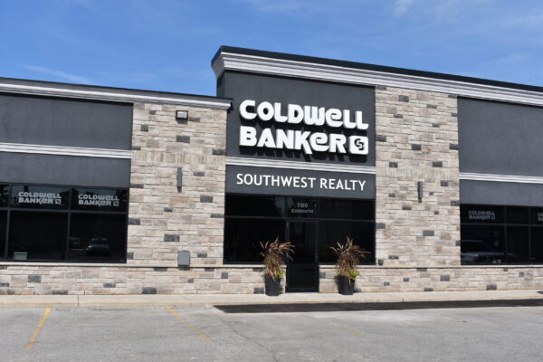 Front view of the Coldwell Banker Southwest Realty building. One-story commercial building with stone and grey brickwork, tinted windows throughout, and on-site parking in front. The building has a white Coldwell Banker sign on the front and a smaller Southwest Realty sign below that.