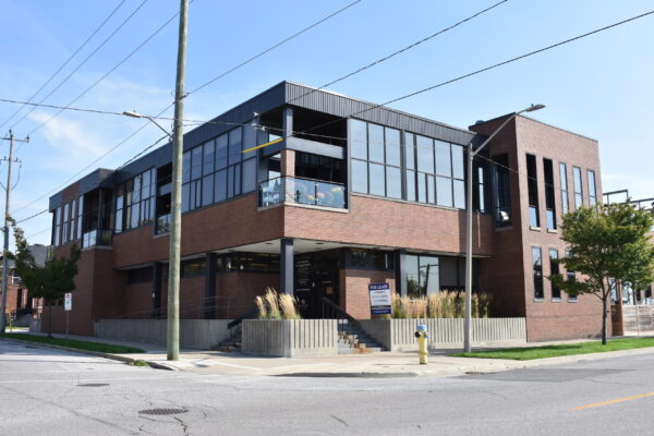 The Sarnia Observer building's exterior view from Front Street in Sarnia. Two stories, brown brick and black siding, tall wall-to-wall windows, a ground level deck and rooftop patios, landscaping with ornamental grasses and trees on the boulevard.
