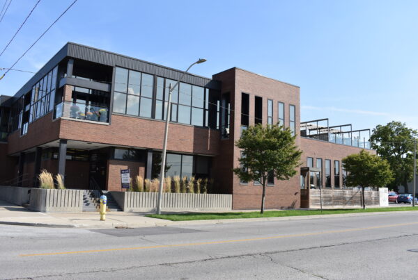The Sarnia Observer building's exterior view. Two stories, brown brick and black siding, tall wall-to-wall windows, a ground level deck and rooftop patios, landscaping with ornamental grasses and trees on the boulevard.