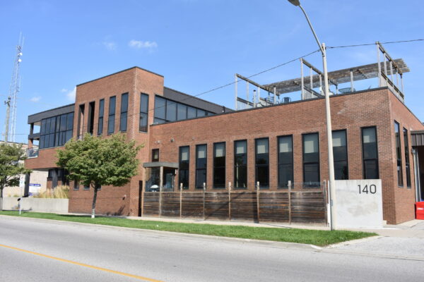 The Sarnia Observer building's exterior view. Two stories, brown brick and black siding, tall wall-to-wall windows, a ground level deck and rooftop patios, landscaping with ornamental grasses and trees on the boulevard.