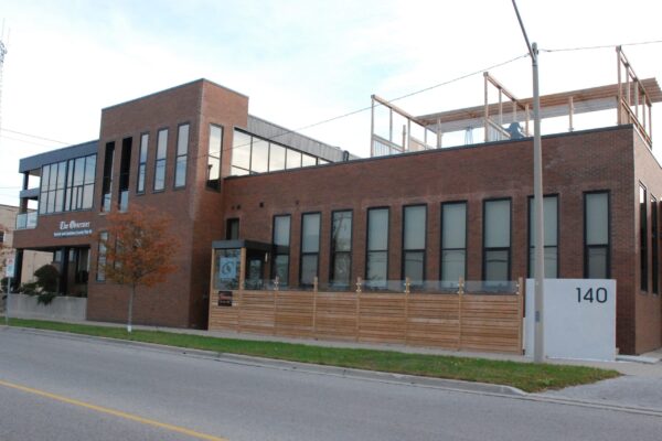 The Sarnia Observer building's exterior view. Two stories, brown brick and black siding, tall wall-to-wall windows, a ground level deck and rooftop patios, landscaping with ornamental grasses and trees on the boulevard.