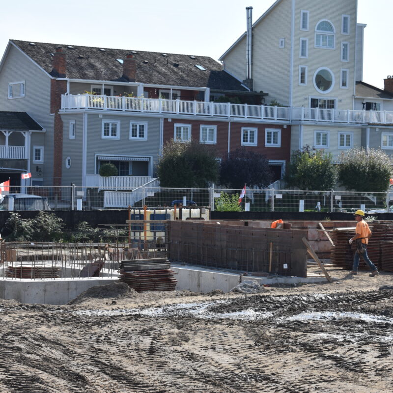 Venetian Boulevard Condominium building in the construction phase. The foreground shows heavy equipment and workers working on foundation for a new building, while another completed building stands in the background, a tall colourful condominium building in blues, yellows, and reds with many large balconies.